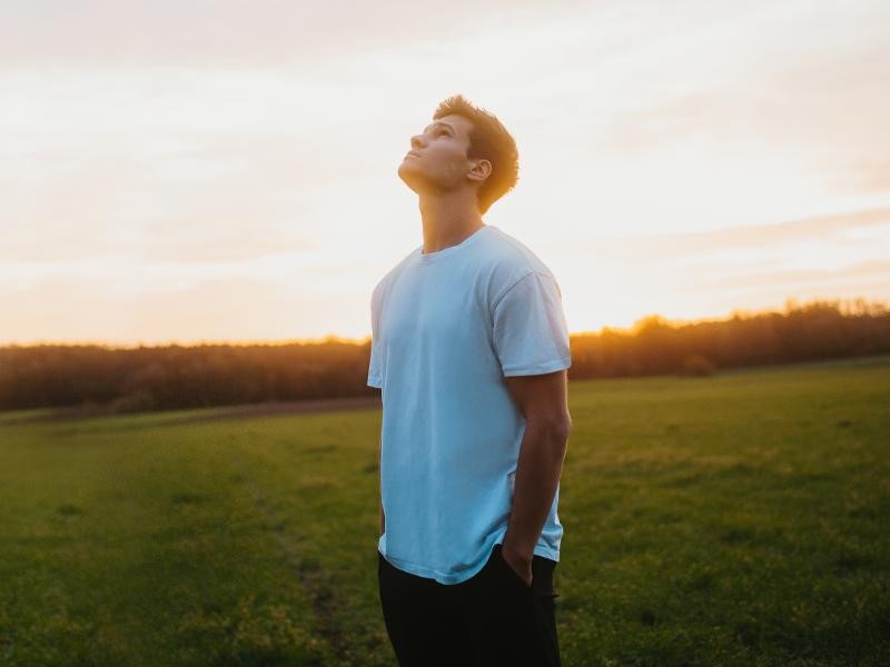 Ein Mann steht auf einer Wiese und schaut in den Himmel. Im Hintergrund sieht man einen Sonnenuntergang.